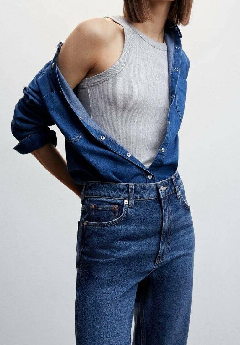 Femme portant un jean bleu taille haute, un top gris sans manches, et une chemise en jean bleue déboutonnée avec les manches retroussées sur un fond uni.