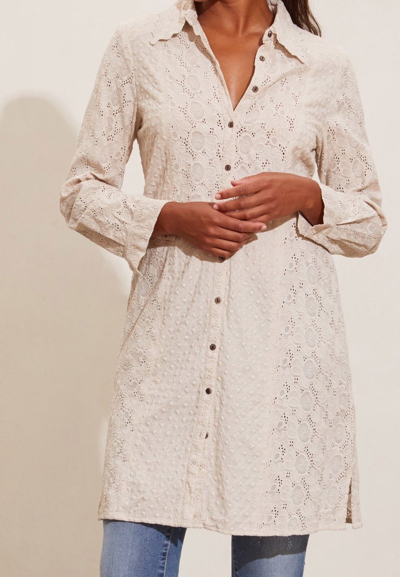 Abito camicia button-up beige chiaro con intricata texture in pizzo, dotato di colletto, maniche lunghe e spacchi laterali. Accenti di bottoni visibili.