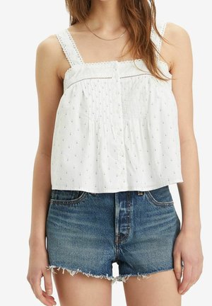 Femme portant un haut blanc sans manches boutonné avec un bord en dentelle et de petits motifs, associé à un short en jean bleu effiloché.