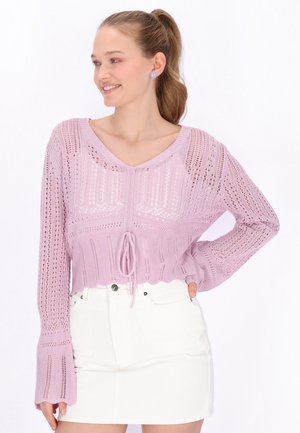 Młoda kobieta uśmiechająca się, ubrana w lawendowy, dziergany sweter z wiązaniem oraz białą, mini spódnicę z wysokim stanem, włosy związane w kucyk.