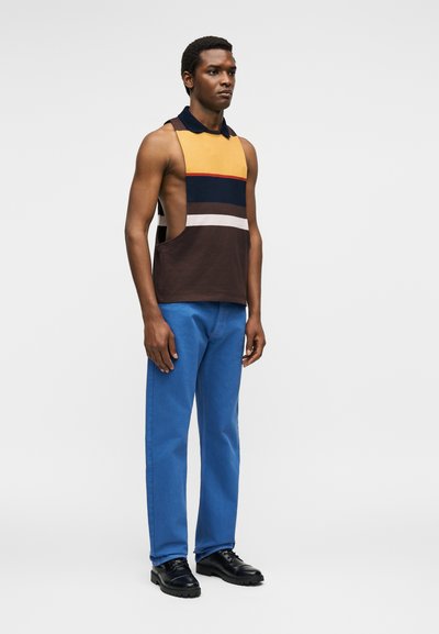 Άνδρας όρθιος με μπλουζάκι χωρίς μανίκια σε μπλοκ χρωμάτων με βαθιά μανικιούλια, μπλε παντελόνι και μαύρα παπούτσια σε απλό φόντο.