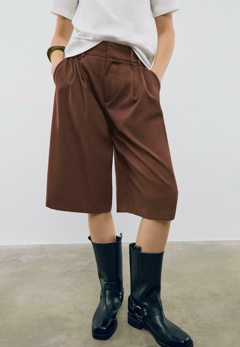 Pantaloni corti marroni su misura con design plissettato, dotati di tasche laterali, abbinati a stivali neri alti in pelle su uno sfondo neutro.