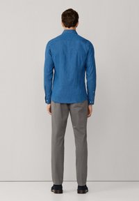 Modrá džínsová košeľa s dlhým rukávom a zapínaním na gombíky, kombinovaná so svetlo šedými nohavicami a tmavými topánkami, pohľad zozadu.