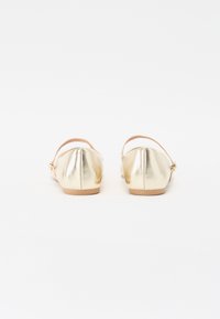 Sapatos planos metálicos dourados com bico arredondado, tira no tornozelo e um detalhe de costura subtil na parte de trás. Textura suave e design minimalista.