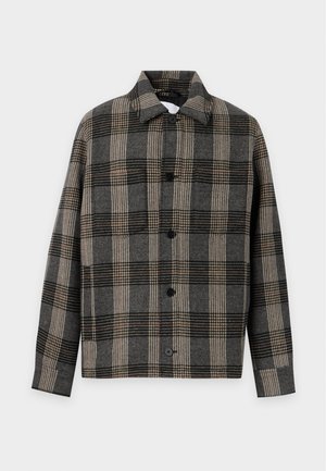 Homme debout portant une veste à carreaux beige et noire à boutons, une chemise blanche, un pantalon noir et des chaussures en cuir noires sur un fond uni.