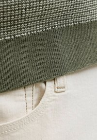 Κοντινό πλάνο στο τελείωμα πουλόβερ ελιάς πάνω από τσέπη μπεζ παντελονιού και ραφές θηλιών ζώνης.