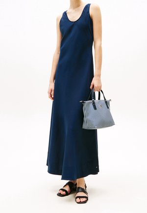 Femme portant une robe maxi bleu marine sans manches, tenant un sac à main rayé, et portant des sandales plates noires sur un fond blanc.