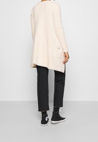 Béžový huňatý svetr na zapínání s dlouhými rukávy a zaobleným spodním lemováním, s jednou boční kapsou ozdobenou dekorativním knoflíkem. Černé džíny a tenisky.