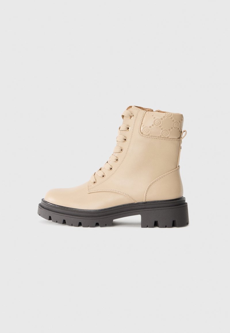 Botte de combat beige à lacets avec cheville rembourrée présentant un motif en relief, semelle épaisse noire à crampons et fermeture éclair latérale sur fond blanc.