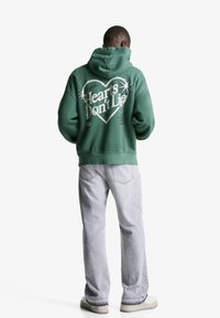 Zelena kapuca s kapuljačom, velika bela grafična napisa "Srca ne lažejo" in zvezde. V kombinaciji s svetlo sivo modrimi hlačami in off-white supergami.