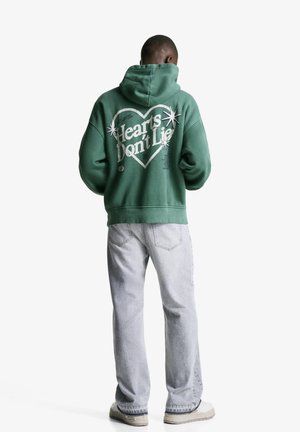 Groene hoodie met kap, grote witte grafische tekst "Hearts Don't Lie" en sterren. Gecombineerd met lichtgrijze jeans en off-white sneakers.
