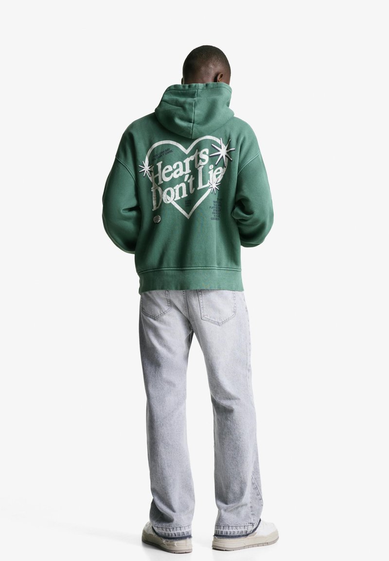 Zelena kapuca s kapuljačom, velika bela grafična napisa "Srca ne lažejo" in zvezde. V kombinaciji s svetlo sivo modrimi hlačami in off-white supergami.