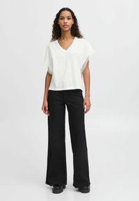 White V-neck top with wide sleeves featuring lace accents, paired with high-waisted black wide-leg trousers and chunky black shoes.