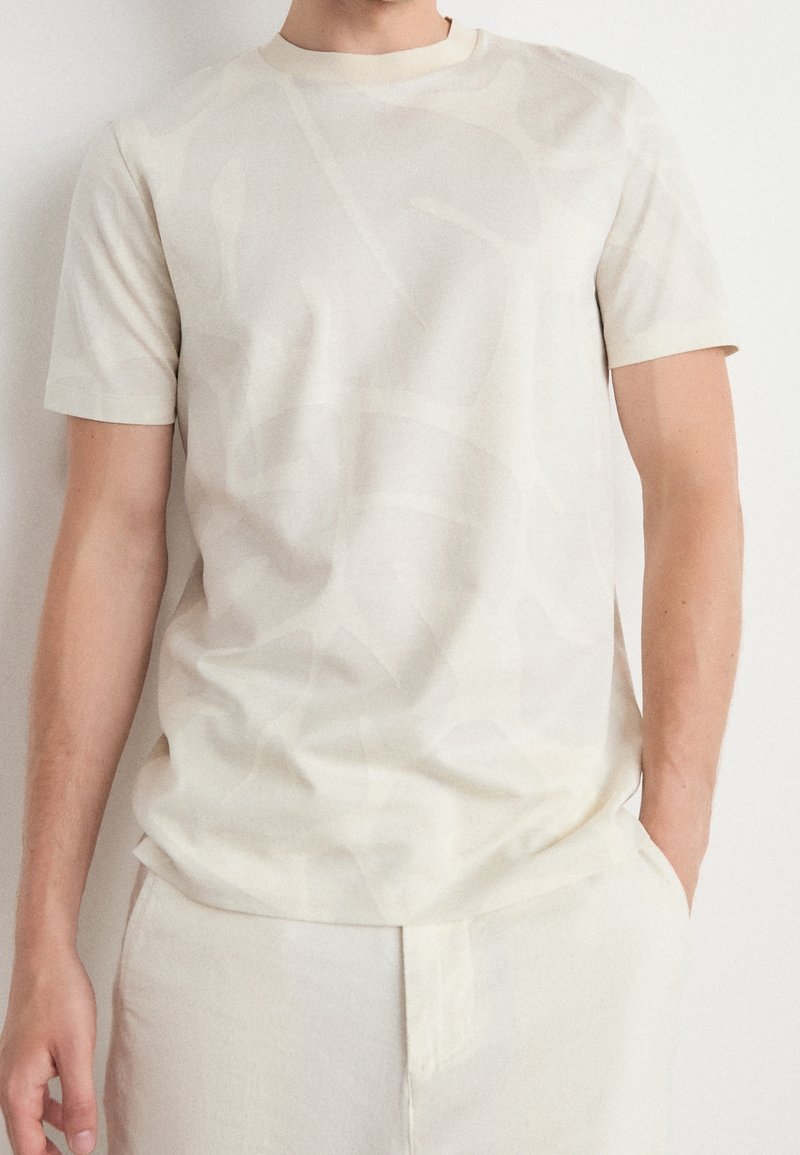 En man bär en off-white kortärmad skjorta och matchande byxor, står mot en enfärgad vit bakgrund med en hand i fickan.