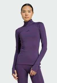 Haut de sport violet à manches longues avec un col montant, texture lisse, orifices pour les pouces et embellissement logo discret sur la poitrine. Design ajusté.