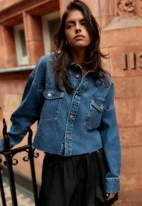 Giacca cropped in denim azzurro chiaro con due tasche sul petto, chiusura a bottoni e colletto a punta, abbinata a una gonna nera.