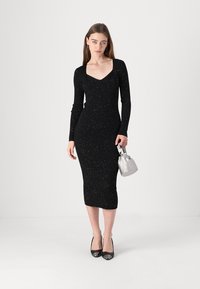 Black ribbed knit dress with long sleeves, V-neckline, mid-calf length, accented with silver speckles. Model holds a small silver handbag.