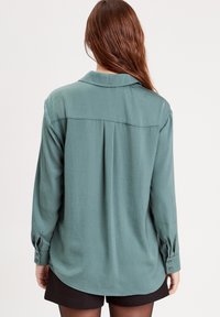 Chemise à manches longues en tissu teal doux avec un col, détail de pli à l'arrière et poignets boutonnés, associée à un short noir.