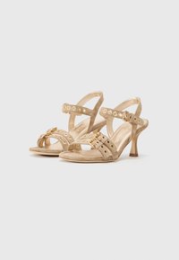 Beige Wildledersandalen mit Absatz, gekreuzten Riemen, goldenen Akzenten und runden Ausschnitten. Mit einem schmalen Stiletto-Absatz und einem strukturierten Fußbett.