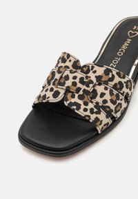 Leopardų rašto šlepetės su smėlio spalvos audiniu ir juodu odiniu padu. Yra kryžminiai diržai ir plokščias dizainas. Viduje auksinis logotipas.