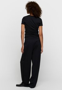 Mujer con cabello rizado de espaldas, vistiendo una camiseta negra ajustada, pantalones negros sueltos y mocasines negros sobre un fondo blanco.