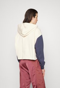 Felpa corta in crema e navy con cappuccio regolabile; abbinata a pantaloni bordeaux lucidi. Tessuto liscio, design casual.
