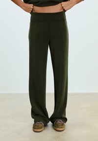 Pantaloni a gamba larga verde oliva con una vita alta e liscia e cuciture verticali. Abbinati a scarpe in suede verdi con lacci a fantasia.
