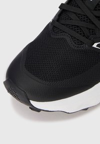 Chaussure de sport noire avec une tige en maille texturée, un embout en caoutchouc et une semelle contrastante blanche. Présente des lacets noirs et des détails de marque.