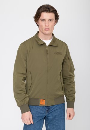 Jonge man draagt een olijfgroene bomberjack met ritssluiting aan de voorkant en een zak op de mouw, gecombineerd met een blauwe spijkerbroek, staand tegen een witte achtergrond.