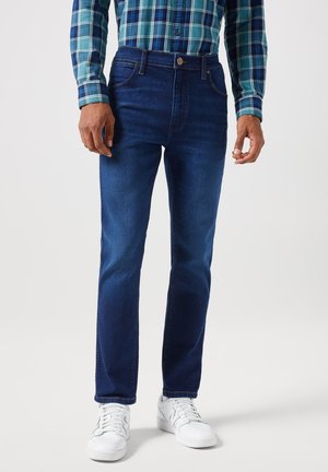 Jeans de mezclilla azul de corte ajustado, diseño de cinco bolsillos y costuras en contraste, combinados con zapatillas blancas y una camisa de cuadros verde.
