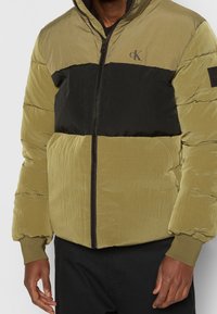 Puffjacka i olivgrönt och svart. Texturerad tyg med dragkedja fram, ribbade muddar och logodetalj på bröstet. Horisontella färgblock.