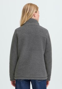 Blond kvinne iført en grå fleecejakke med høy krage, sett fra bak, kombinert med blå denimjeans.