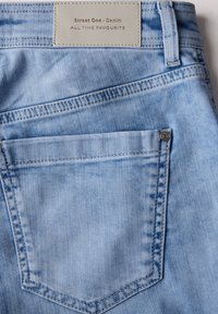 Ljust blå denimjeans med en sydd bakficka och en beige etikett med texten "Street One - Denim". Mjuk textur med blekningsdetaljer.