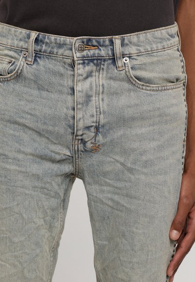 Wyblakłe niebieskie dżinsy z zapięciem na guzik, pięcioma kieszeniami i kontrastowym szwem. Powierzchnia ma postrzępioną teksturę i zagięcia.