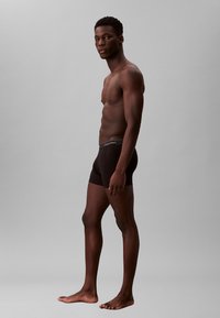 Schwarze Boxershorts aus weichem Stoff, mit einem Bund, der das Logo von Calvin Klein zeigt, dargestellt an einem Modell im Seitenprofil vor einem grauen Hintergrund.