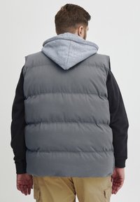 Grå polstret vest med hette. Vesten har et quiltet design, en glatt tekstur og armhull. Under er en svart langermet skjorte synlig.
