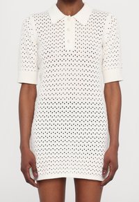 Robe tricotée blanche avec col polo, manches courtes et un design texturé ouvert comportant des pois noirs. Fermeture à trois boutons au niveau de la poitrine.