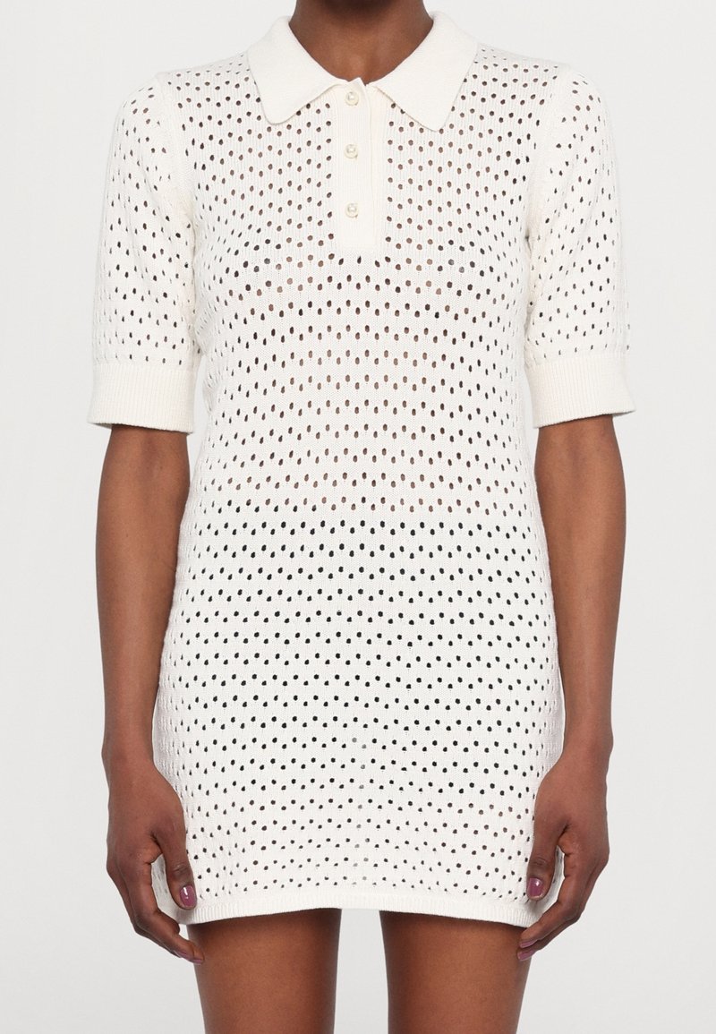 Robe tricotée blanche avec col polo, manches courtes et un design texturé ouvert comportant des pois noirs. Fermeture à trois boutons au niveau de la poitrine.