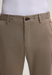 Beige broeken met een gladde textuur, voorzien van een platte tailleband, knoopsluiting en schuine zakken. Subtiel visgraatpatroon zichtbaar.