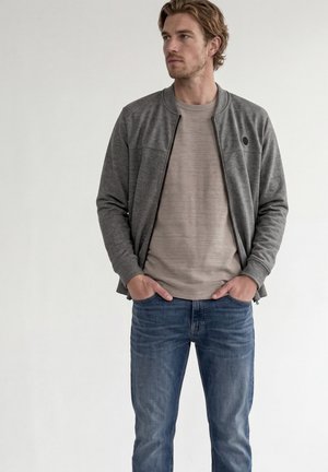 Man met een grijze jas met rits over een beige overhemd en blauwe spijkerbroek, staand met handen in de zakken, kijkend opzij tegen een effen achtergrond.