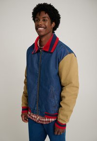 Denim jacket with brown sleeves, red and blue striped collar, front zipper, and ribbed cuffs. Worn over a plaid shirt and blue jeans.