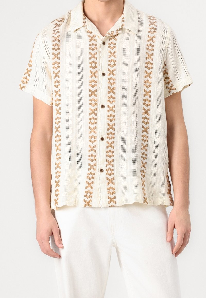 Homme portant une chemise à manches courtes boutonnée avec des motifs croisés beige verticaux et un pantalon blanc sur un fond uni.