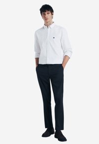 Camicia bianca con colletto, caratterizzata da un logo blu. Abbinata a pantaloni scuri e scarpe nere, creando un look elegante e formale.