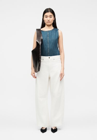 Γυναίκα με αμάνικο τζιν τοπ, άσπρο παντελόνι με φαρδύ πόδι, μαύρα μυτερά παπούτσια και κρατώντας μεγάλη μαύρη υφασμάτινη τσάντα ώμου.