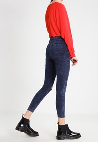 Stivaletti neri in vernice con un leggero tacco, indossati con jeans skinny in denim scuro e una blusa a maniche lunghe rossa intensa. Tessuti lisci e vestibilità curata.