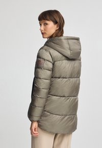 Olivgrön pufferjacka med huva, quiltad textur, sidofickor och en brun logotypapplikation på vänster arm.