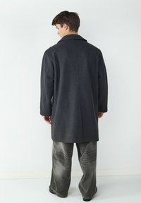 Langer grauer Wollmantel mit weitem Kragen, geradem Schnitt und glatter Textur. Kombiniert mit hellgrauen, weit geschnittenen Jeans.