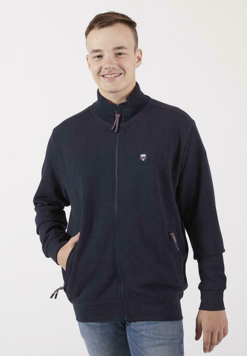 Sweatshirt zippé navy avec un col haut, deux poches latérales et des poignets côtelés. Présente un petit logo sur la poitrine gauche. Texture douce.