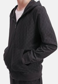 Persona con una sudadera oscura con cremallera y un sutil patrón circular, manos en los bolsillos frontales, y una camiseta blanca debajo, recortada en el cuello.