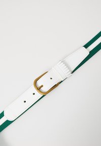 Vihreä kudottu vyö, jossa on valkoinen nahkahihna ja kultainen metallinappi. Nahassa on aaltoileva reuna ja useita säätöreikiä.